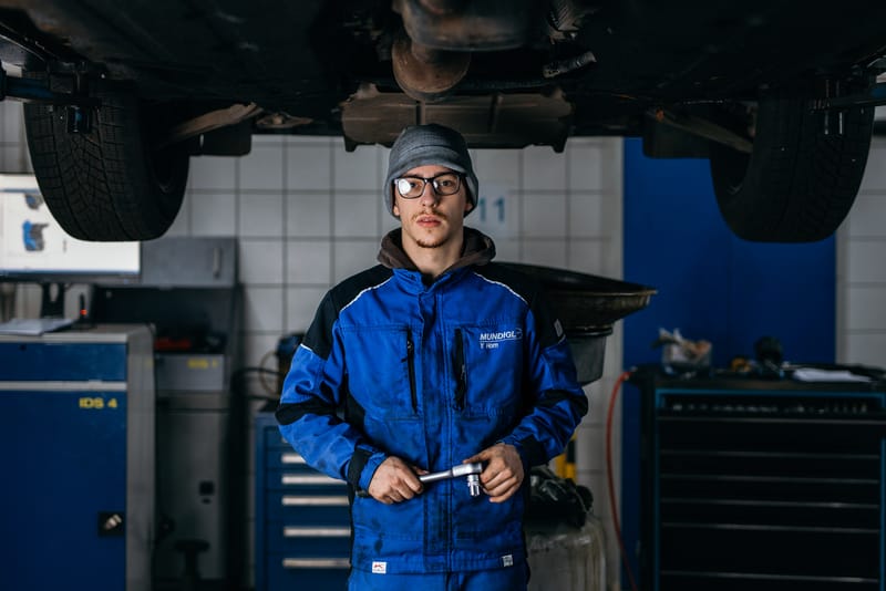 Ein junger Mechaniker steht in Arbeitskleidung unter einem Fahrzeug auf der Hebebühne